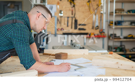 A portrait of a carpenter in goggles and work overalls draws a blueprint for a workshop. 99412704