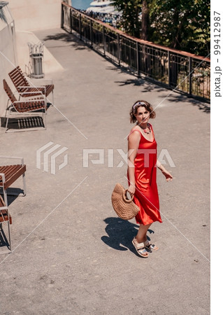 woman in a red silk dress and a bandage on her head smiles against the background of the leaves of a tree. She is leaning on the coop and looking into the camera. Vertical photo. woman in a red silk dress and a bandage on her head smiles against the background of the leaves of a tree. She is leaning on the coop and looking into the camera. Vertical photo. 99412987