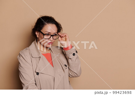 Confident Latin American middle-aged business woman in beige casual coat, putting on stylish specs - eyeglasses, squinting, looking away, standing isolated on beige background. Vision problems concept Confident Latin American middle-aged business woman in beige casual coat, putting on stylish specs - eyeglasses, squinting, looking away, standing isolated on beige background. Vision problems concept 99423698