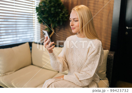 Portrait of attractive young woman sitting on couch, looking at mobile phone screen, reading online content, browsing internet, shopping with application. Happy blonde female browsing smartphone apps 99423732