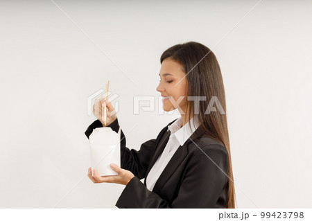 Portrait of happy young woman in business suit holding paper box with fast food noodles and bamboo sticks in her hands. Studio shot of attractive brunette girl on isolated white background. Fast food 99423798