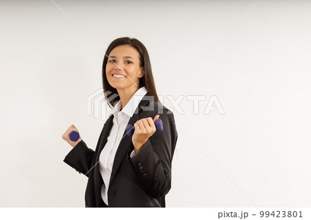 Attractive girl with dark hair in stylish business suit and white shirt holds small female dumbbells. Portrait of happy smiling young woman with sports equipment on isolated white background. Attractive girl with dark hair in stylish business suit and white shirt holds small female dumbbells. Portrait of happy smiling young woman with sports equipment on isolated white background. 99423801