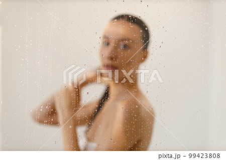 Young woman under the trickles of water in pleasure shower. Beautiful girl under the spray of water. Wet through glass drops selective focus Young woman under the trickles of water in pleasure shower. Beautiful girl under the spray of water. Wet through glass drops selective focus 99423808