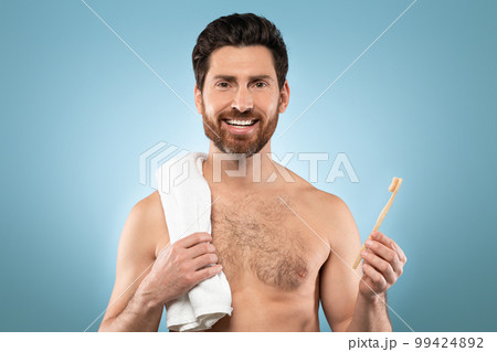 Portrait of handsome bearded man with towel on shoulder smiling at camera, standing with toothbrush over blue background 99424892