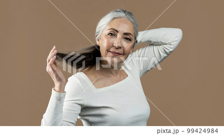Cheerful retired european female touching gray hair with hands, looking at camera, isolated on brown background 99424984