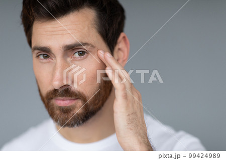 Anti-aging eye care treatment. Handsome middle aged man touching under eyes skin, closeup, grey background 99424989