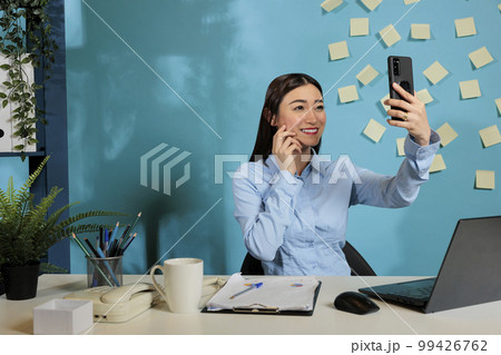 Corporate asian female employee sitting at contemporary office desk taking selfie with smart phone. Professional woman smiling in front of mobile device screen at work station. 99426762