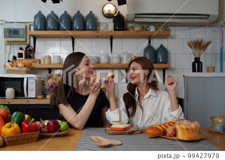 Beautiful woman and friend talking and using laptop at kitchen room. Girl friends couple living together at home. 99427978