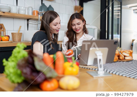 Beautiful woman and friend talking and using laptop at kitchen room. Girl friends couple living together at home. 99427979