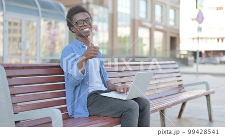 Young African Man with Laptop Showing Thumbs Up Sign While Sitting Outdoor on Bench 99428541