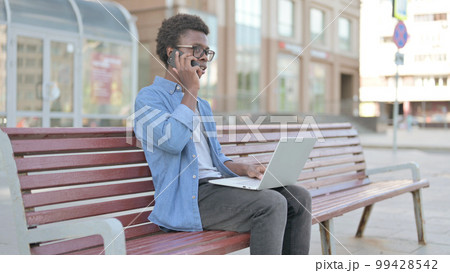 Young African Man Talking on Phone and using Laptop while Sitting Outdoor on Bench Young African Man Talking on Phone and using Laptop while Sitting Outdoor on Bench 99428542