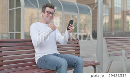 Young Man Celebrating Online Success on Smartphone while Sitting Outdoor on Bench Young Man Celebrating Online Success on Smartphone while Sitting Outdoor on Bench 99428555