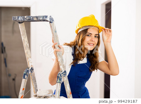 Portrait of contented young female builder in blue overalls during the repair of cottage 99429187