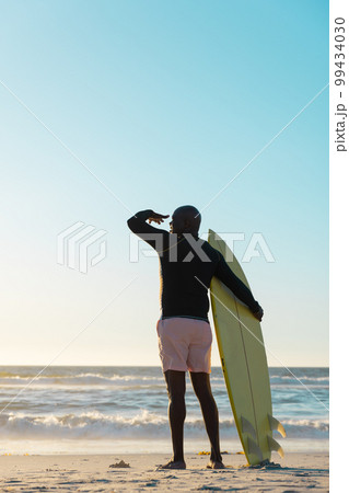 Rear view of senior african american man with surfboard looking at sea under clear sky at sunset Rear view of senior african american man with surfboard looking at sea under clear sky at sunset 99434030