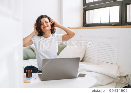 Young brunette woman with closed eyes in headphones working on laptop from home Young brunette woman with closed eyes in headphones working on laptop from home 99434602