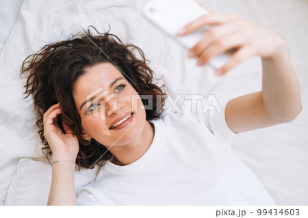 Young smiling brunette woman in home clothes using mobile on bed at home, top view 99434603