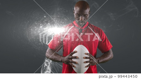 African american male rugby player holding ball against smoke and light spot on grey background African american male rugby player holding ball against smoke and light spot on grey background 99443450