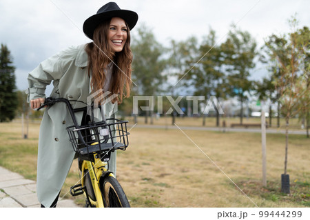 cheerful young woman in a gray coat and black hat rides a bike with a smile 99444299