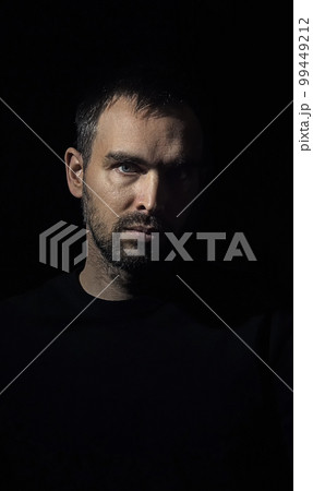 Portrait of a stern man with a small beard looking into the camera. Caucasian man on dark background. Half of his face illuminated. Dramatic portrait of a handsome man. Portrait of a stern man with a small beard looking into the camera. Caucasian man on dark background. Half of his face illuminated. Dramatic portrait of a handsome man. 99449212