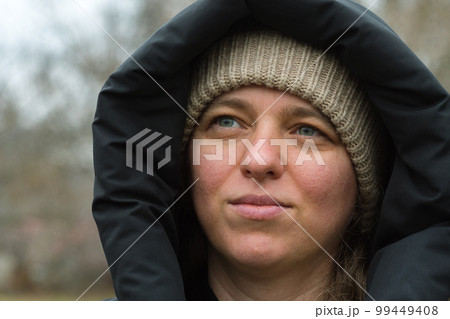 A close-up of the face of a Caucasian girl wearing a knitted cap and hood. The woman's gaze is dreamily turned to the side. Beautiful eyes. Cold weather. Out of season. A close-up of the face of a Caucasian girl wearing a knitted cap and hood. The woman's gaze is dreamily turned to the side. Beautiful eyes. Cold weather. Out of season. 99449408
