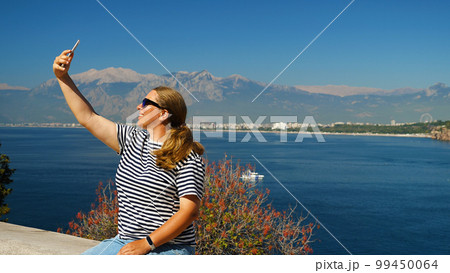 Cheerful attractive girl takes selfie portrait on smartphone. Woman in sunglasses takes selfie on phone while on vacation. Beautiful landscape with light mountains and blue sea in the background. Cheerful attractive girl takes selfie portrait on smartphone. Woman in sunglasses takes selfie on phone while on vacation. Beautiful landscape with light mountains and blue sea in the background. 99450064