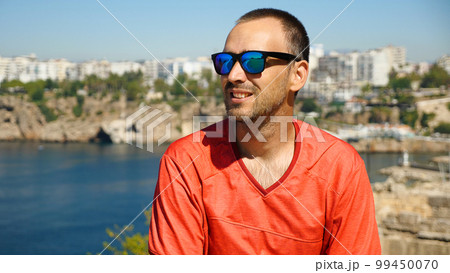 Handsome man with sunglasses smiling against a background of sea and city. Caucasian man in red jersey against a beautiful seascape with mountains. 99450070