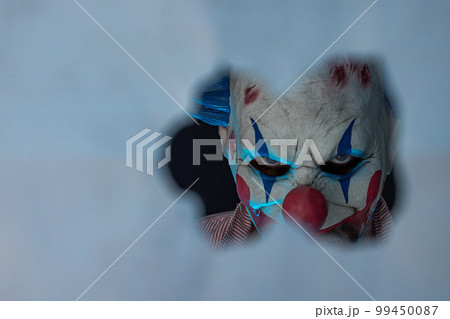 Close-up of a creepy clown looking through a hole. The face of a man wearing a creepy clown mask, looking at his victim. Blue lighting of the crazy clown. Close-up of a creepy clown looking through a hole. The face of a man wearing a creepy clown mask, looking at his victim. Blue lighting of the crazy clown. 99450087