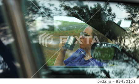 A girl with glasses is drinking her morning coffee. View through the window of a car parked at a campsite. Woman drinking a hot drink from a mug while sitting in a car at a campsite. A girl with glasses is drinking her morning coffee. View through the window of a car parked at a campsite. Woman drinking a hot drink from a mug while sitting in a car at a campsite. 99450153
