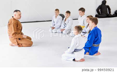 Group of little boys, children in kimono sitting on floor around coach. Children training indoors. Judo, jiu-jitsu training Group of little boys, children in kimono sitting on floor around coach. Children training indoors. Judo, jiu-jitsu training 99450921
