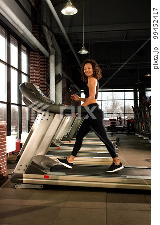 Smiling beautiful African American woman running on a treadmill in the gym. Weight loss, cardio training, keeping muscles in good shape. 99452417