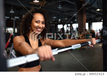 Beautiful young smiling african american woman looking at metal disc on barbell before hard bench press workout. Close-up. 99452423