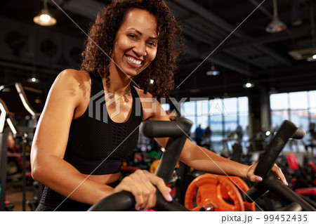 African American smiling woman doing cardio workout on exercise bike in gym smiling at camera. 99452435