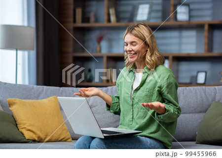 Cheerful and happy mature woman alone at home sitting on sofa in living room and using laptop for video call, housewife smiling and talking with friends remotely. 99455966