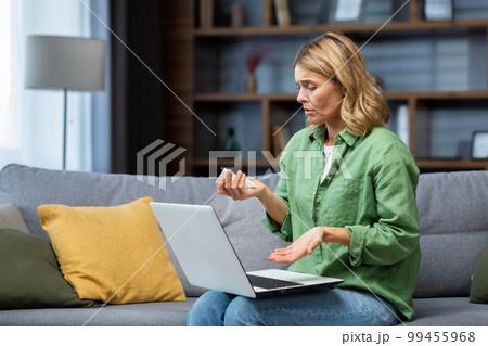 Mature lonely woman alone at home, crying in depression talking on video call with psychologist, sitting on sofa in living room, using laptop for online psychological support. 99455968