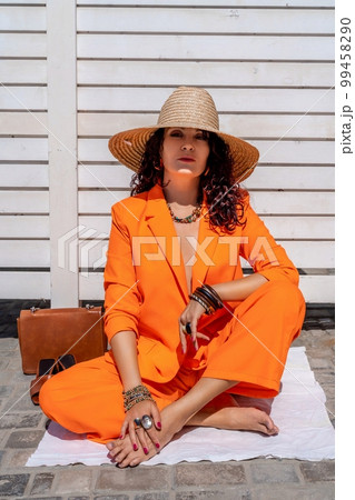 Stylish woman in an orange suit with a hat sits on a rug on a white striped background. On the hands are jewelry rings and bracelets, sandals and a bag stand side by side. 99458290