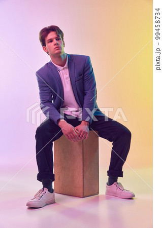 Handsome caucasian young man, business man in jacket and jeans sitting on wooden box over light gradient yellow-purple background. Handsome caucasian young man, business man in jacket and jeans sitting on wooden box over light gradient yellow-purple background. 99458734