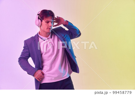 Half-length portrait of young man in headphones dancing isolated on gradient yellow-purple background in neon light. Concept of human emotions, youth culture. Half-length portrait of young man in headphones dancing isolated on gradient yellow-purple background in neon light. Concept of human emotions, youth culture. 99458779