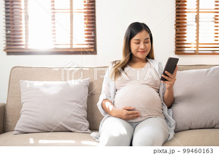 Happy Asian pregnant woman using her phone while relaxing on sofa in her living room Happy Asian pregnant woman using her phone while relaxing on sofa in her living room 99460163