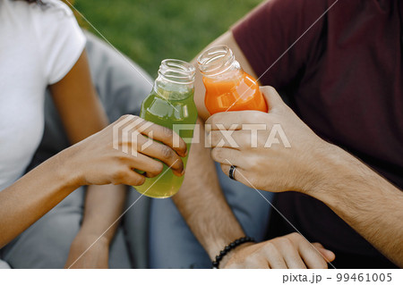 Focus on a bottles. Friends hanging in the green park, relaxing, sitting on the bean bag chairs on grass. Boy and girl clinking a bottles of juice. Focus on a bottles. Friends hanging in the green park, relaxing, sitting on the bean bag chairs on grass. Boy and girl clinking a bottles of juice. 99461005