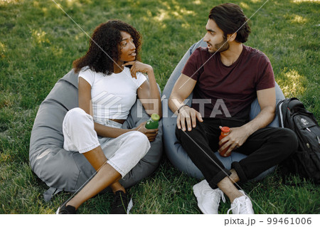 Friends hanging in the green park, relaxing, sitting on the bean bag chairs on grass. Boy and girl holding a bottles of juice. African girl wearing white clothes, boy wearing brown t-shirt and black Friends hanging in the green park, relaxing, sitting on the bean bag chairs on grass. Boy and girl holding a bottles of juice. African girl wearing white clothes, boy wearing brown t-shirt and black 99461006