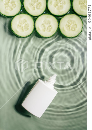 White beauty product cream vial tube with row of cucumber slices on surface of rippled transparent green water. Vertical White beauty product cream vial tube with row of cucumber slices on surface of rippled transparent green water. Vertical 99461771