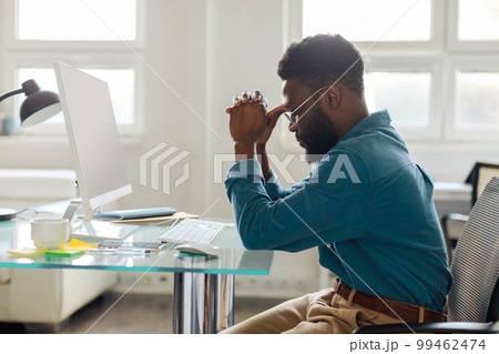 Thoughtful black male entrepreneur in glasses sitting at desk in front of computer, working online in office, side view Thoughtful black male entrepreneur in glasses sitting at desk in front of computer, working online in office, side view 99462474