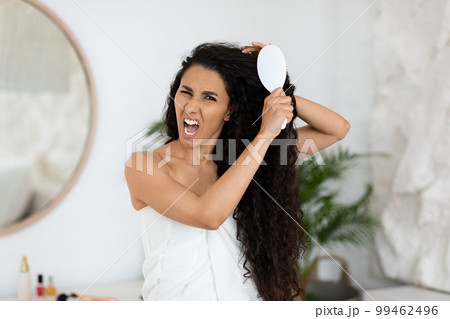 Angry sad millennial caucasian brunette lady in towel combing long curly hair, suffer from pain and tangled Angry sad millennial caucasian brunette lady in towel combing long curly hair, suffer from pain and tangled 99462496