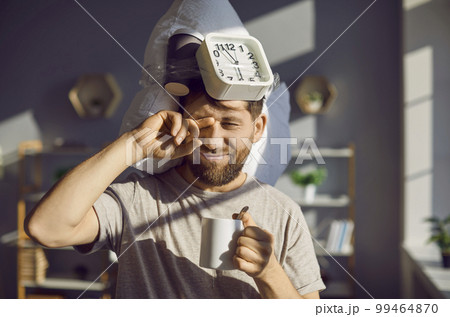 Man with a pillow and an alarm clock drinking morning coffee and rubbing his eyes 99464870