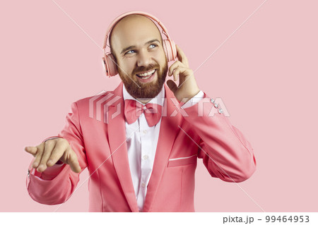 Young man in pink party suit, bow tie and headphones listening to music and smiling Young man in pink party suit, bow tie and headphones listening to music and smiling 99464953