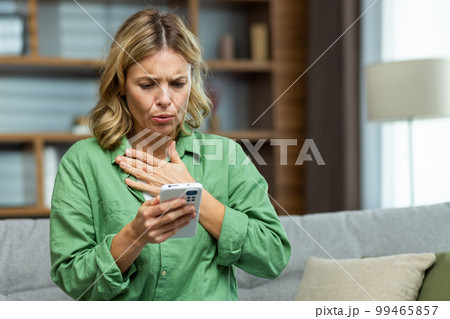 Lonely mature woman alone close up holding phone upset and depressed reading bad news on smartphone online in living room close up. 99465857