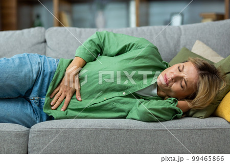 Mature woman alone at home sick, holding hands to side of stomach having severe spasm lying on sofa in living room. Mature woman alone at home sick, holding hands to side of stomach having severe spasm lying on sofa in living room. 99465866