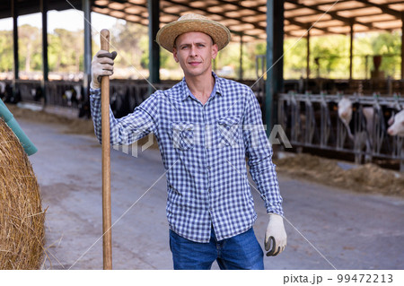Male farmer holding agricultural tool and standing in hangar 99472213