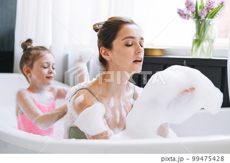 Young mother woman and little tween girl daughter having fun in the bath with foam at home 99475428