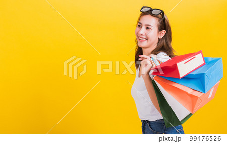 Asian Thai portrait happy beautiful cute young woman smiling stand with sunglasses excited holding shopping bags multi color looking side, studio shot isolated yellow background with copy space 99476526